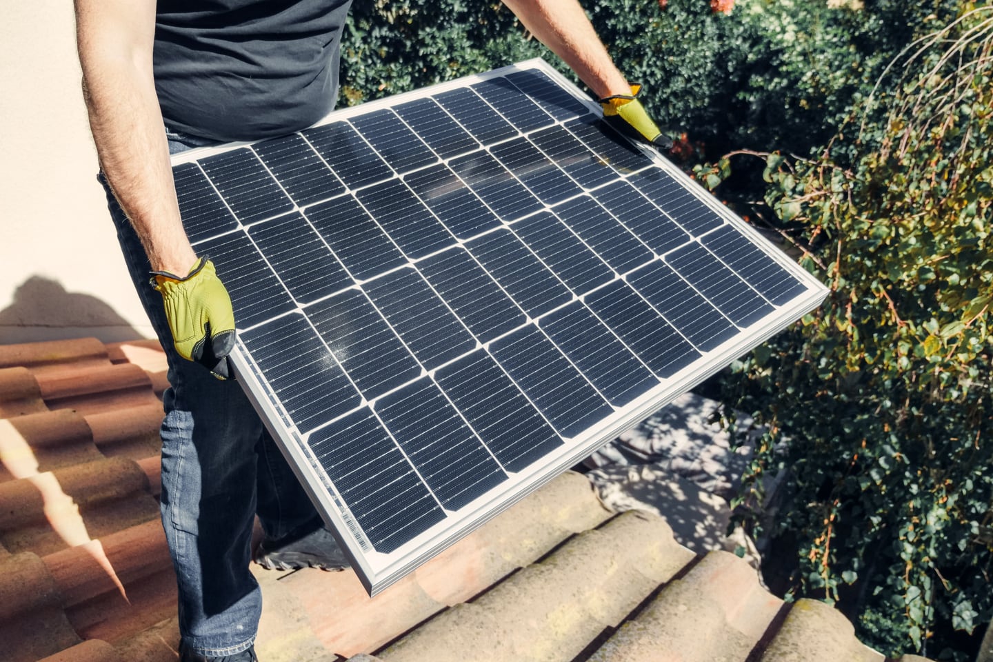 A worker carries a solar panel across a tiled roof.